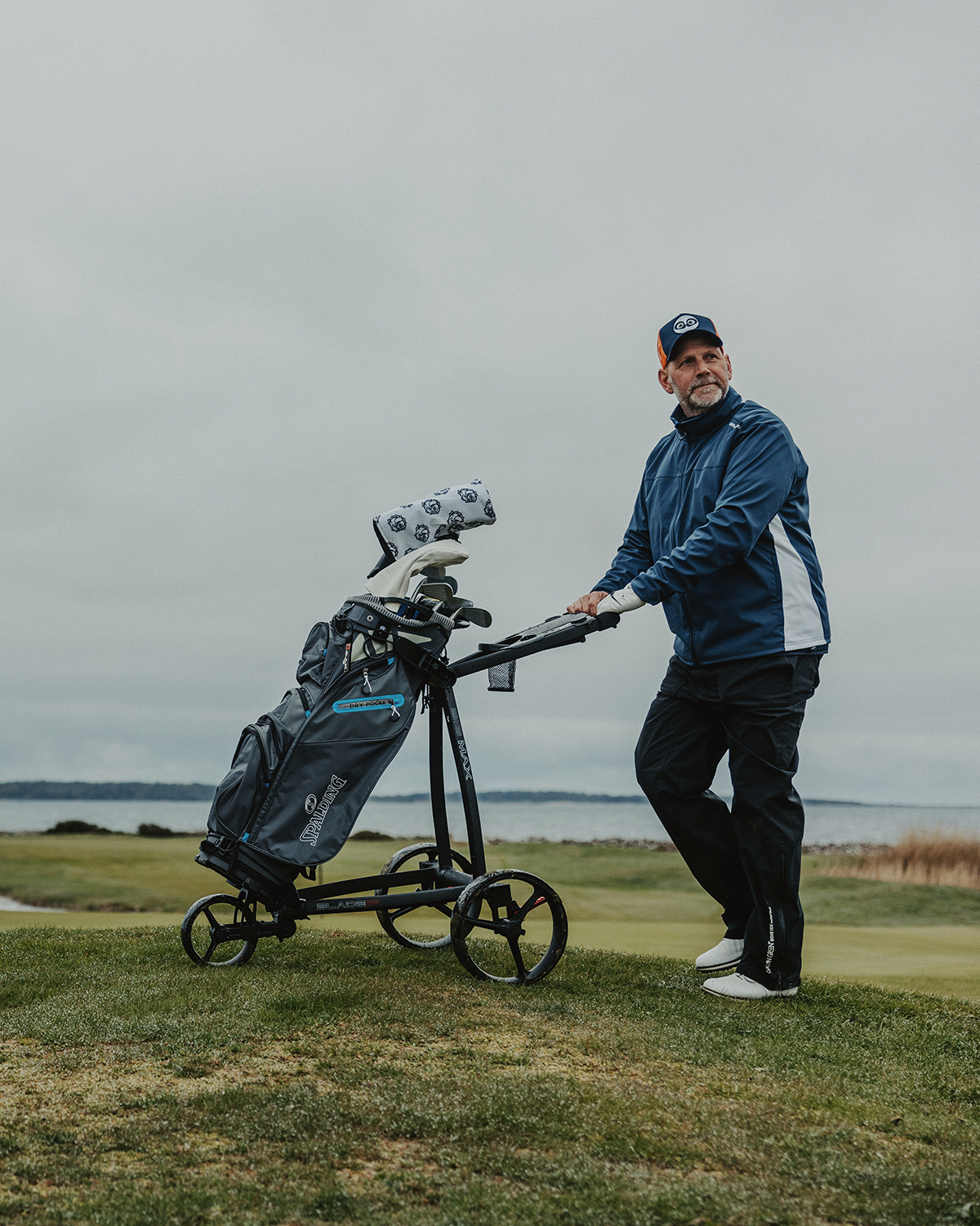 Ein Mann in blauer Jacke und Mütze schiebt einen Golfwagen mit Golfschlägern auf einem grünen Golfplatz in der Nähe des Wassers unter bewölktem Himmel.