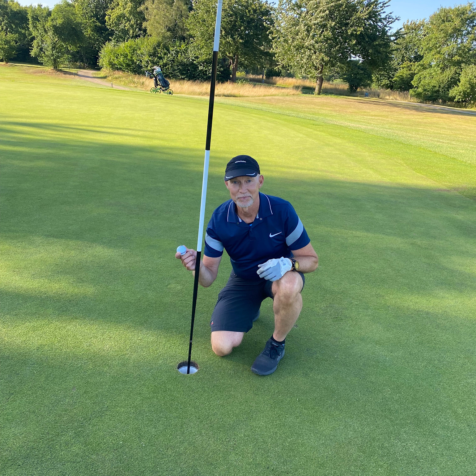 Mand iført golfbeklædning hukker ved hullet på en golfbane, holder en golfbold og poserer ved siden af flagstangen. Grønne omgivelser i baggrunden.