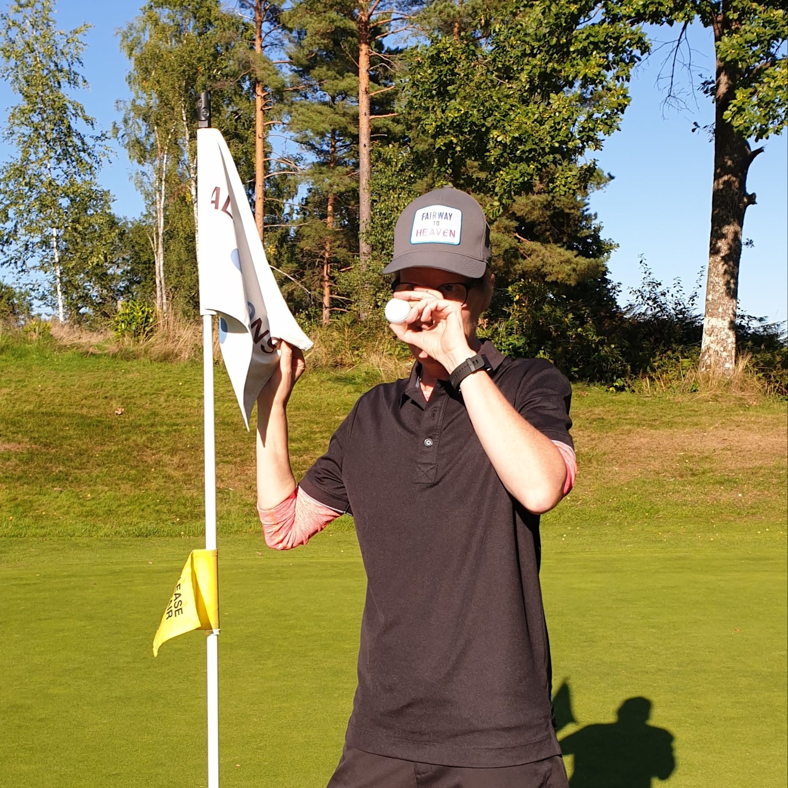 En person holder en golfbold foran øjet og står ved siden af en flagstang på en golfbane på en solrig dag.