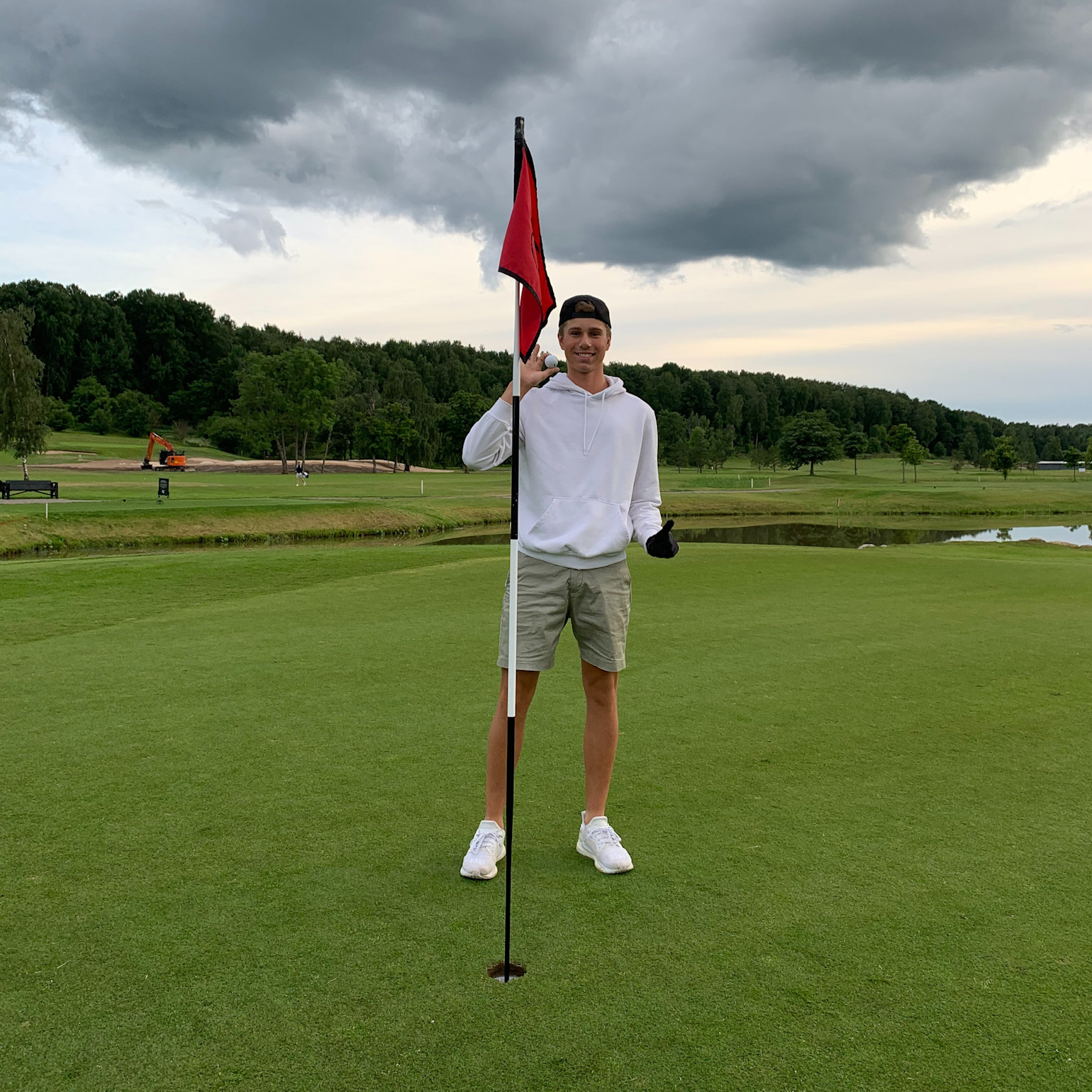 Ung mand i hvid hættetrøje og kasket står ved et golf-flag på en green, omgivet af græs, træer og en overskyet himmel.