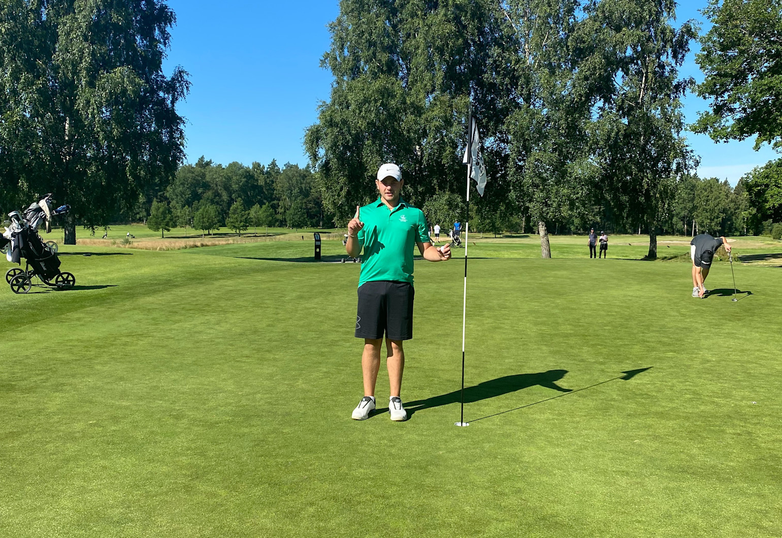 Mand i grøn poloshirt står ved siden af et golfhul og en flag, holder én finger op, på en solrig golfbane med træer i baggrunden.