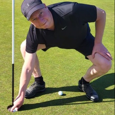 Man i svart sportkläder böjer sig ner för att ta upp en golfboll ur hålet på en golfbana i solsken.