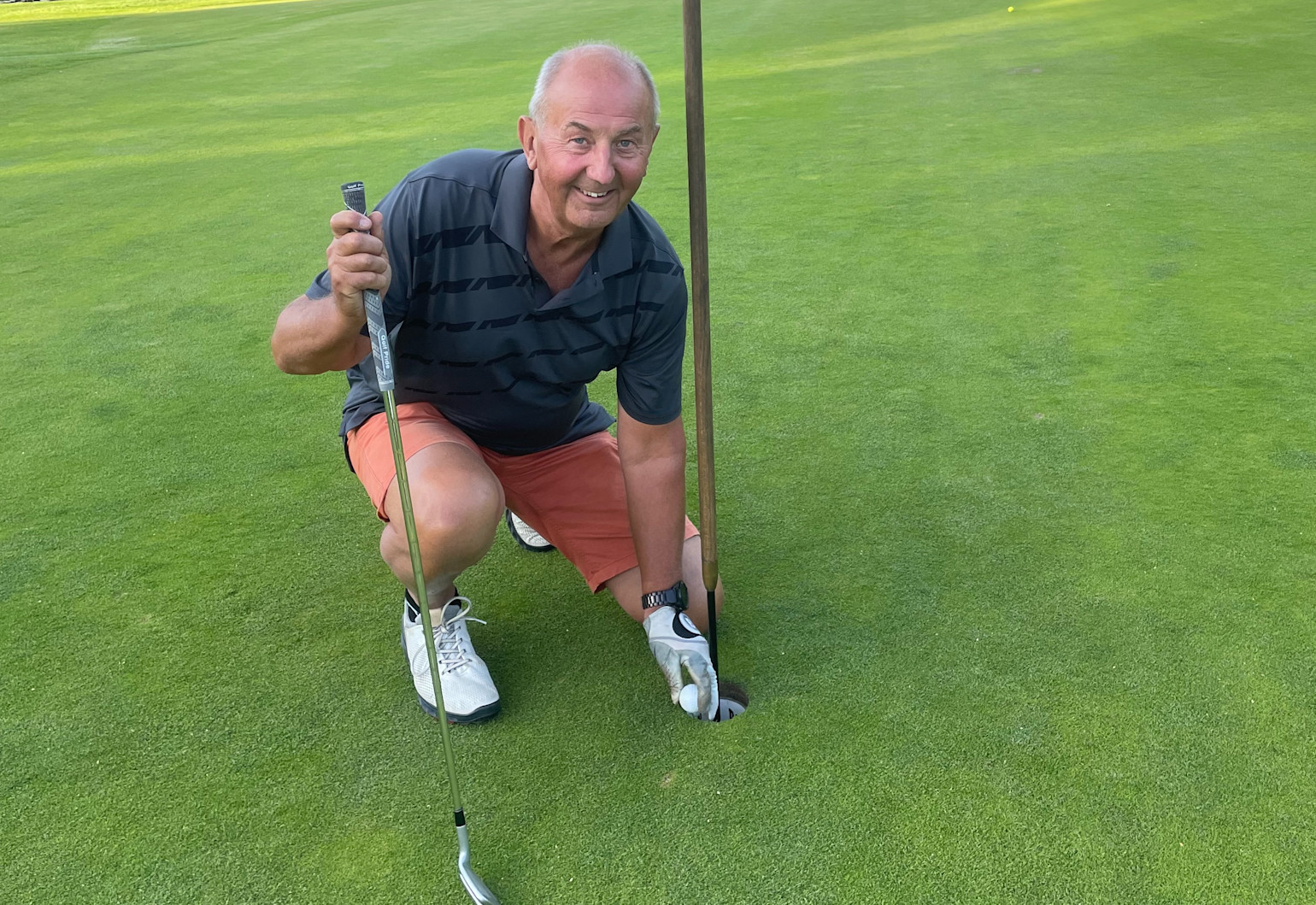 Ældre mand i golftøj samler en golfbold op af hullet på en golfbane og smiler til kameraet.