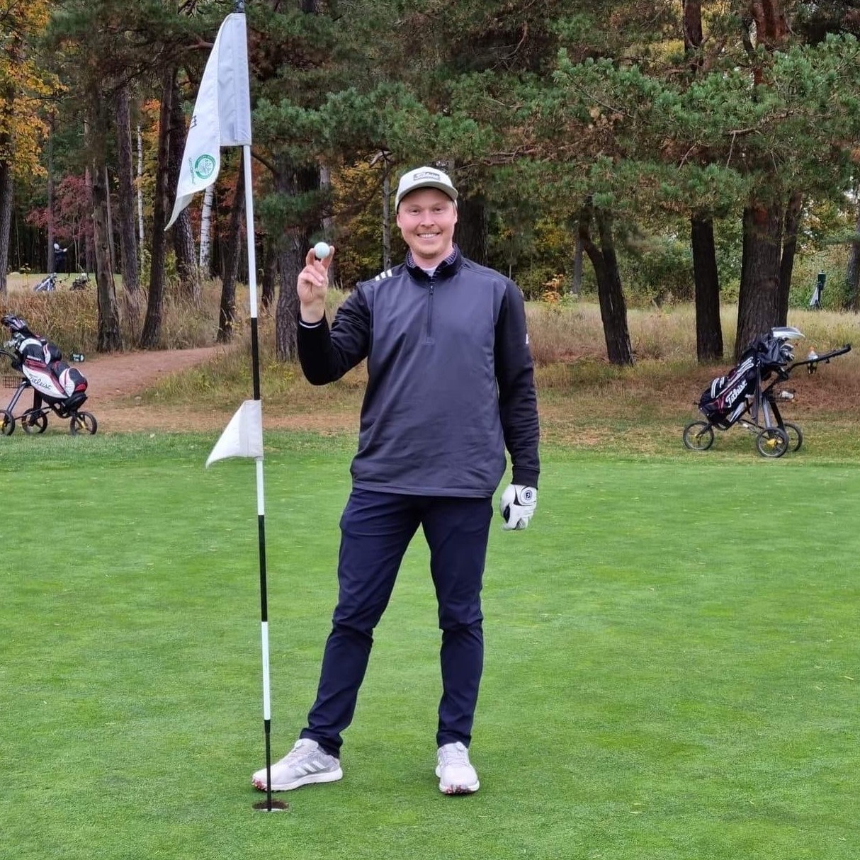 Man smiler og holder en golfbold op ved siden af et hul med flag på en golfbane, omgivet af træer og golfvogne i baggrunden.