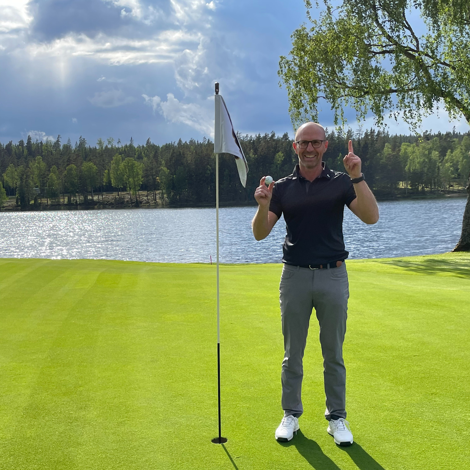 Man smiler og holder en golfbold op ved siden af en flagstang på en golfgreen ved en sø, med skov i baggrunden.