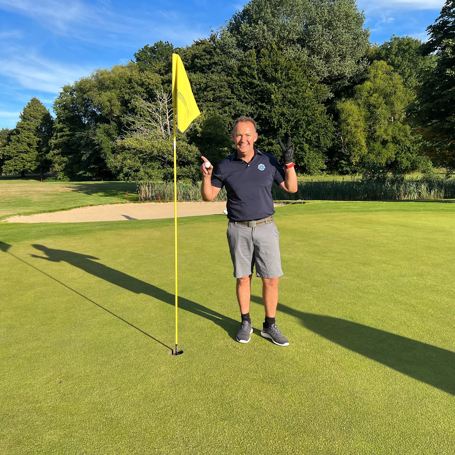 Mand iført golfbeklædning står smilende ved siden af et gult flag på en golfgreen, holder en golfbold og en handske op, med træer i baggrunden.