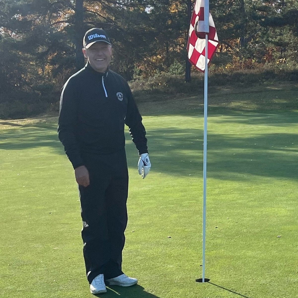 Man i golfkläder står leende bredvid en flaggstång på en golfgreen med träd i bakgrunden.