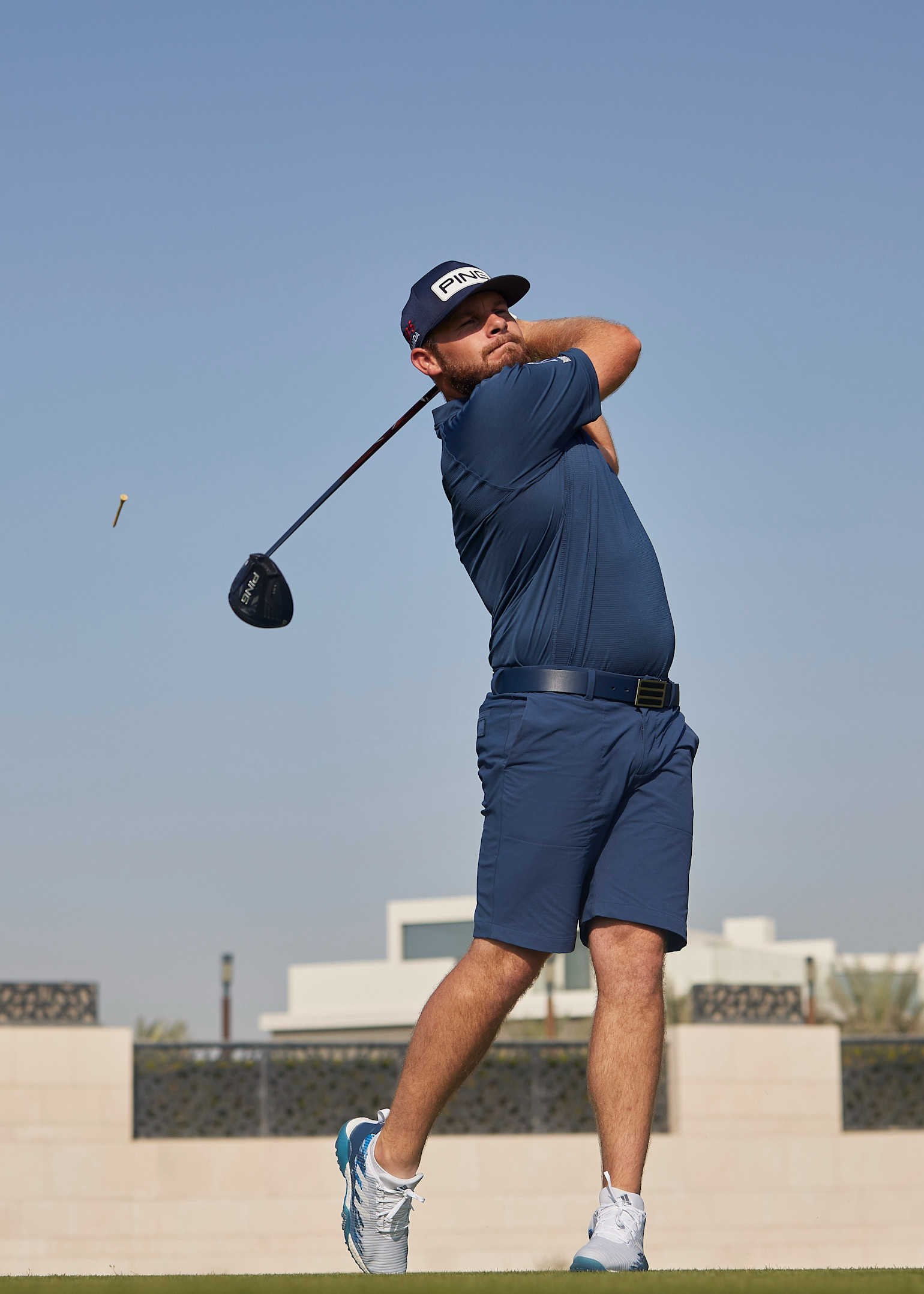 Ein Mann in blauer Golfkleidung schwingt einen Golfschläger im Freien auf einem Golfplatz, mit blauem Himmel im Hintergrund.