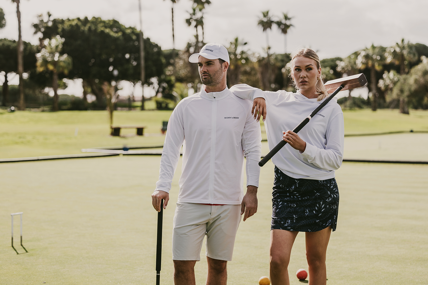 Två personer i sportkläder står på en croquetbana, båda håller croquetklubbor, med träd och gräs i bakgrunden.