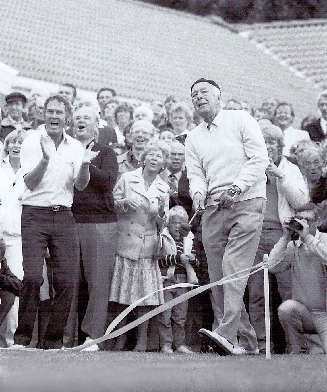 En äldre man slår ett golfslag medan en glad publik applåderar och hejar på honom utomhus på en golfbana.
