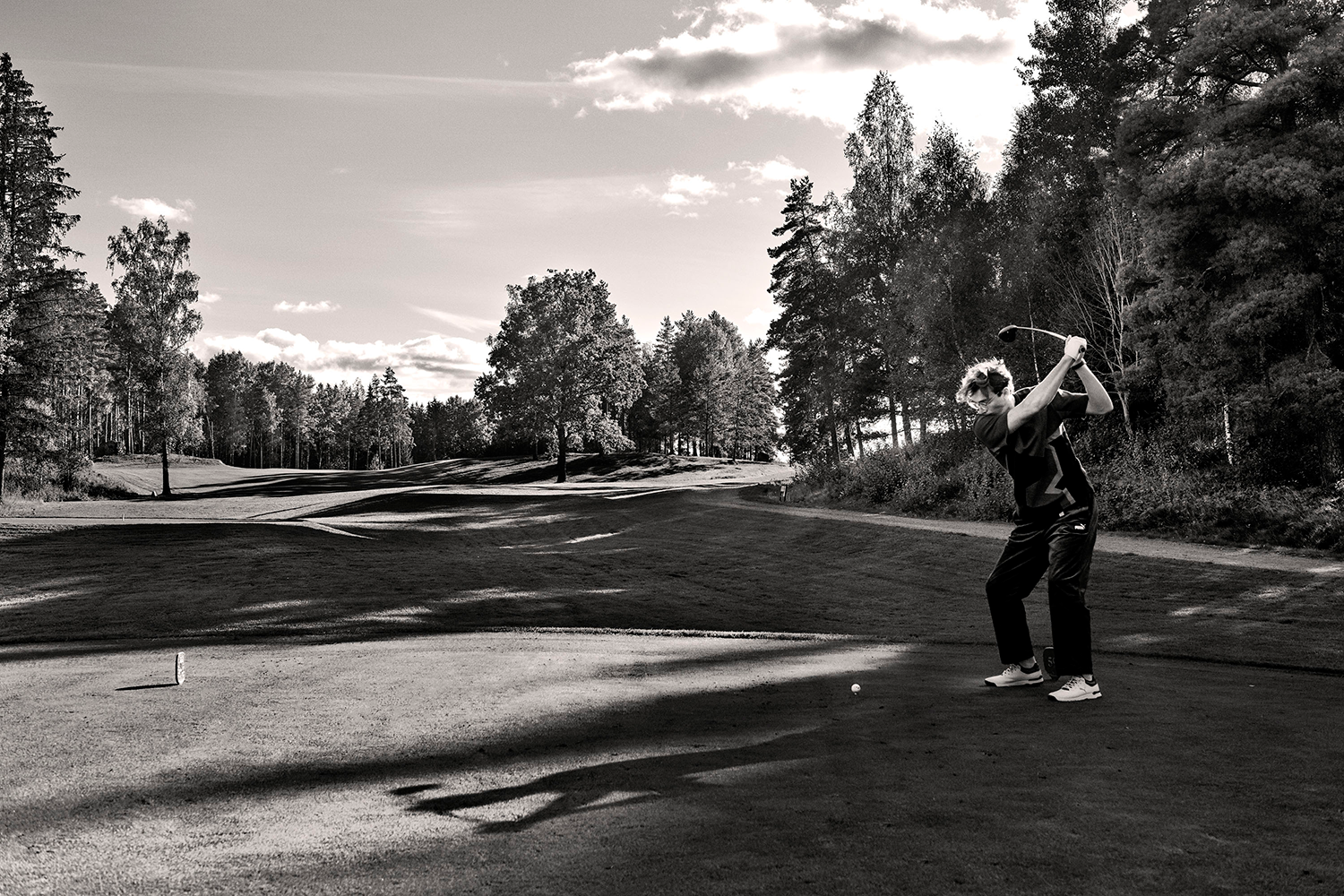 En person svingar en golfklubba på en golfbana omgiven av träd under en solig himmel.