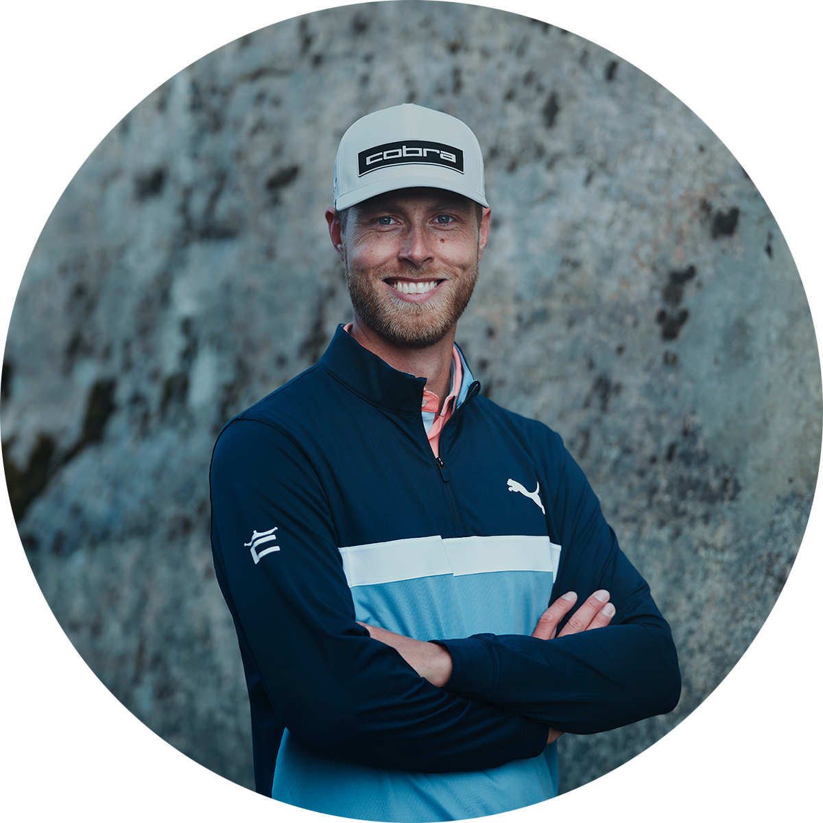 Smiling man wearing a cap with the text "cobra" and a blue sports jacket with a Puma logo, standing with his arms crossed in front of a rocky background.