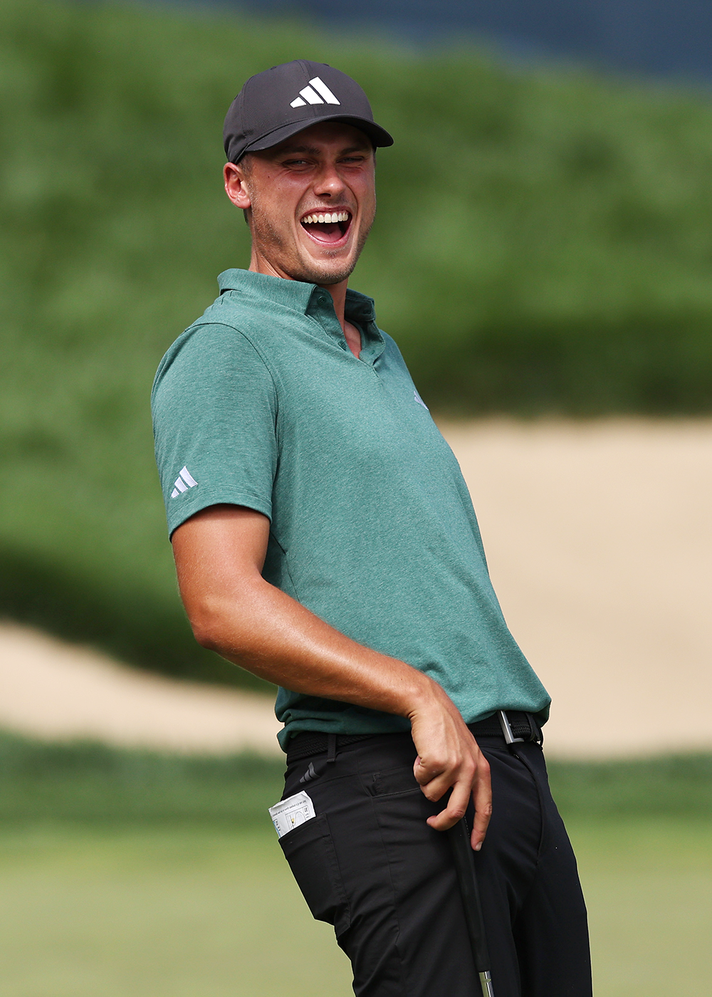 En man i grön pikétröja och svart keps skrattar och håller i en golfklubba på en golfbana.