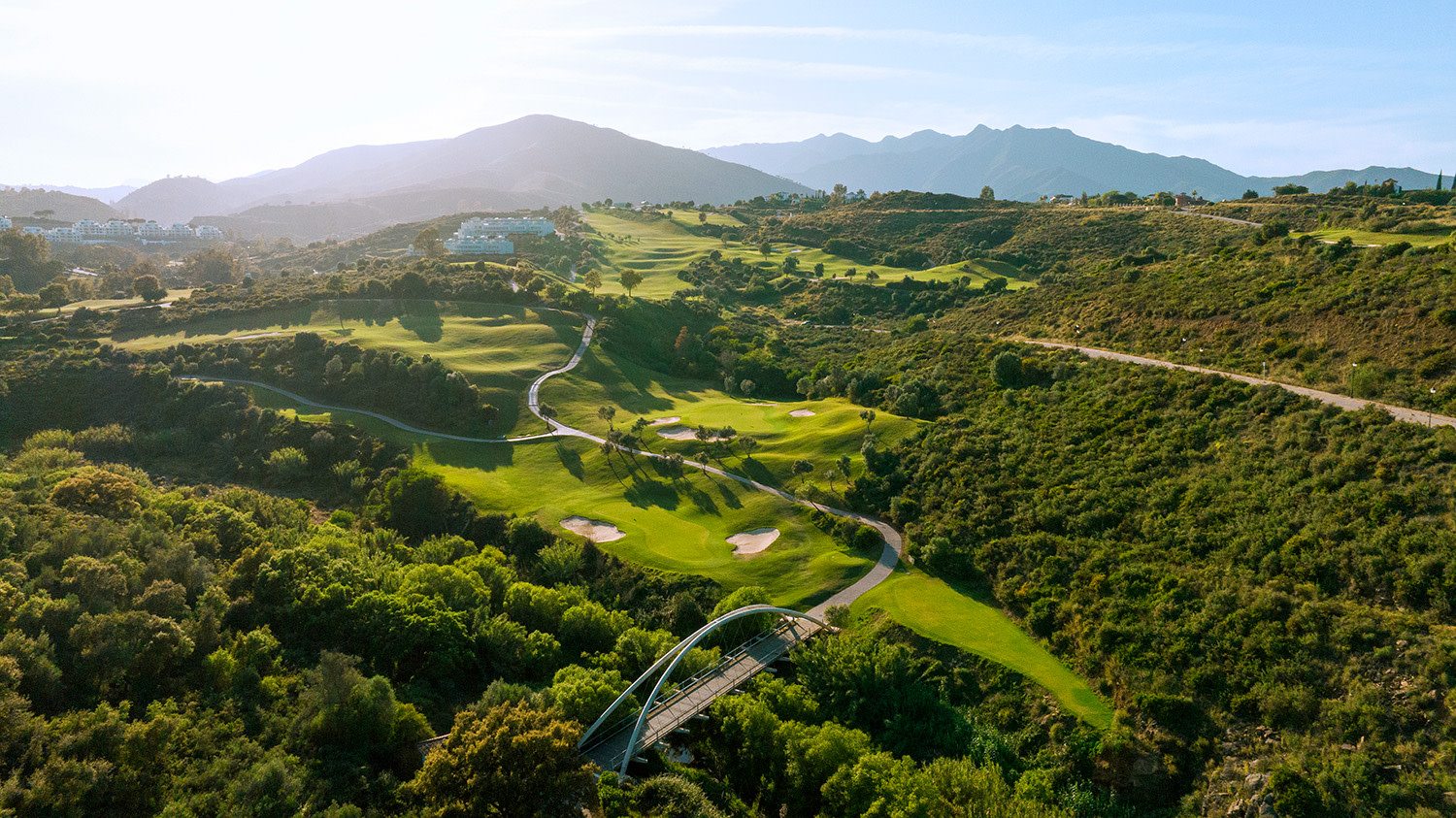 Grön golfbana med sandbunkrar slingrar sig genom kuperat landskap, omgiven av träd och berg i bakgrunden under klar himmel.