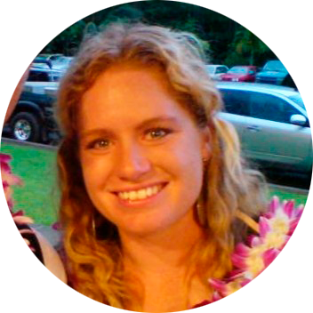 Leende kvinna med lockigt blont hår bär blommor runt halsen, bilar och grönska i bakgrunden.