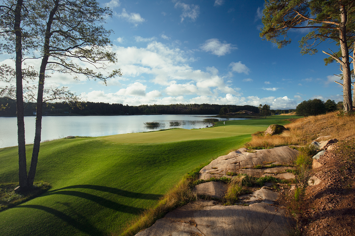 Grön golfbana vid en sjö med flagga på green, omgiven av träd, klippor och blå himmel med moln.