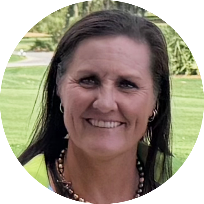 Kvinna med långt mörkt hår ler utomhus på en grön gräsmatta, bär halsband och örhängen.