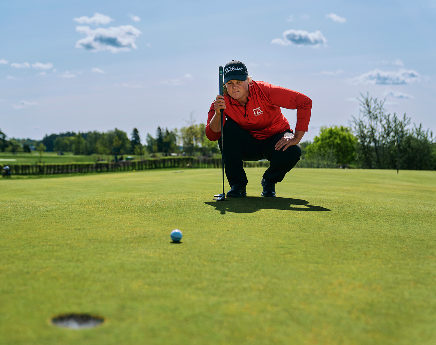 Golfspelare i röd tröja hukar sig och siktar mot ett hål på greenen med en golfklubba, solig dag utomhus.