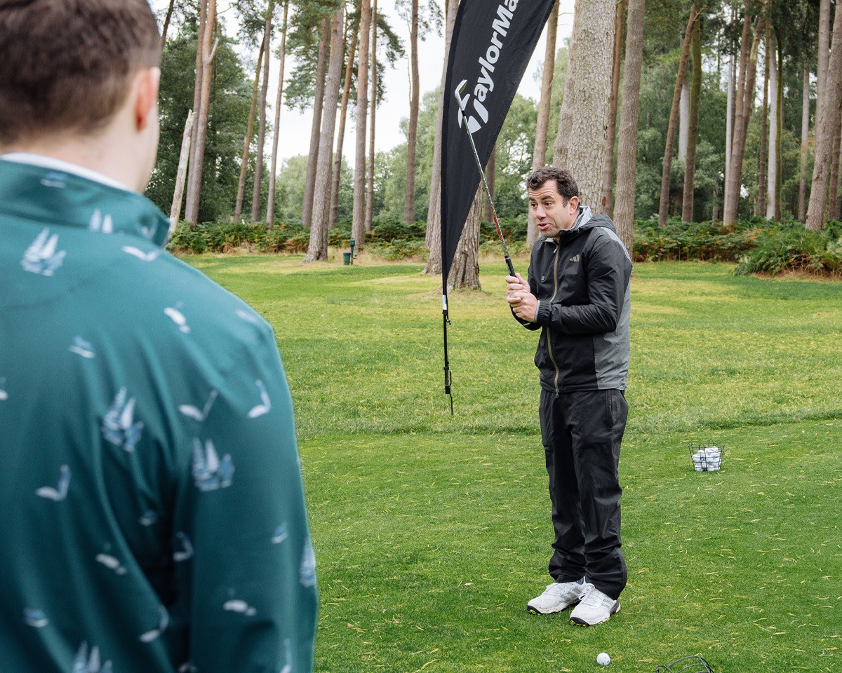 En golftränare visar en svingteknik med en klubba för en elev på en grön golfbana omgiven av träd.