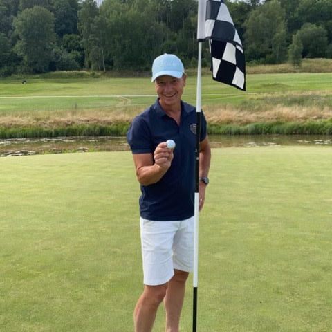 En mand står på en golfgreen, holder en golfbold og smiler ved siden af et sort-hvidt flag, med træer og græs i baggrunden.