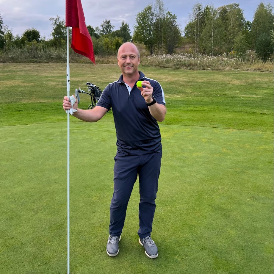 Man holder en golfkølle og en gul golfbold, står ved siden af flaget på en green med en golfbag i baggrunden.