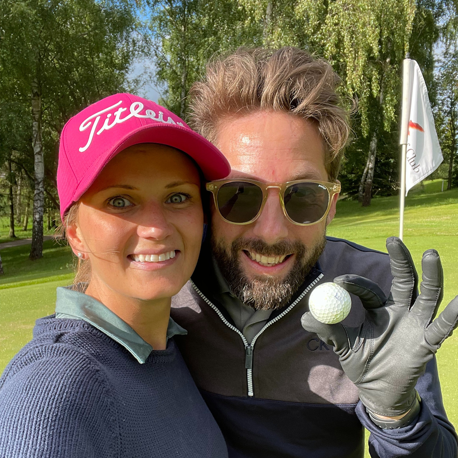 To glade personer poserer på en golfbane, en kvinde med lyserød kasket og en mand med solbriller, som holder en golfbold.