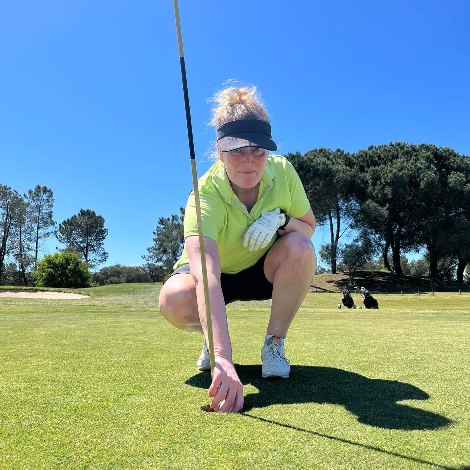 Kvinde i golfbeklædning sætter sig på hug og placerer en golfbold på greenen ved siden af flagstangen, med træer i baggrunden.