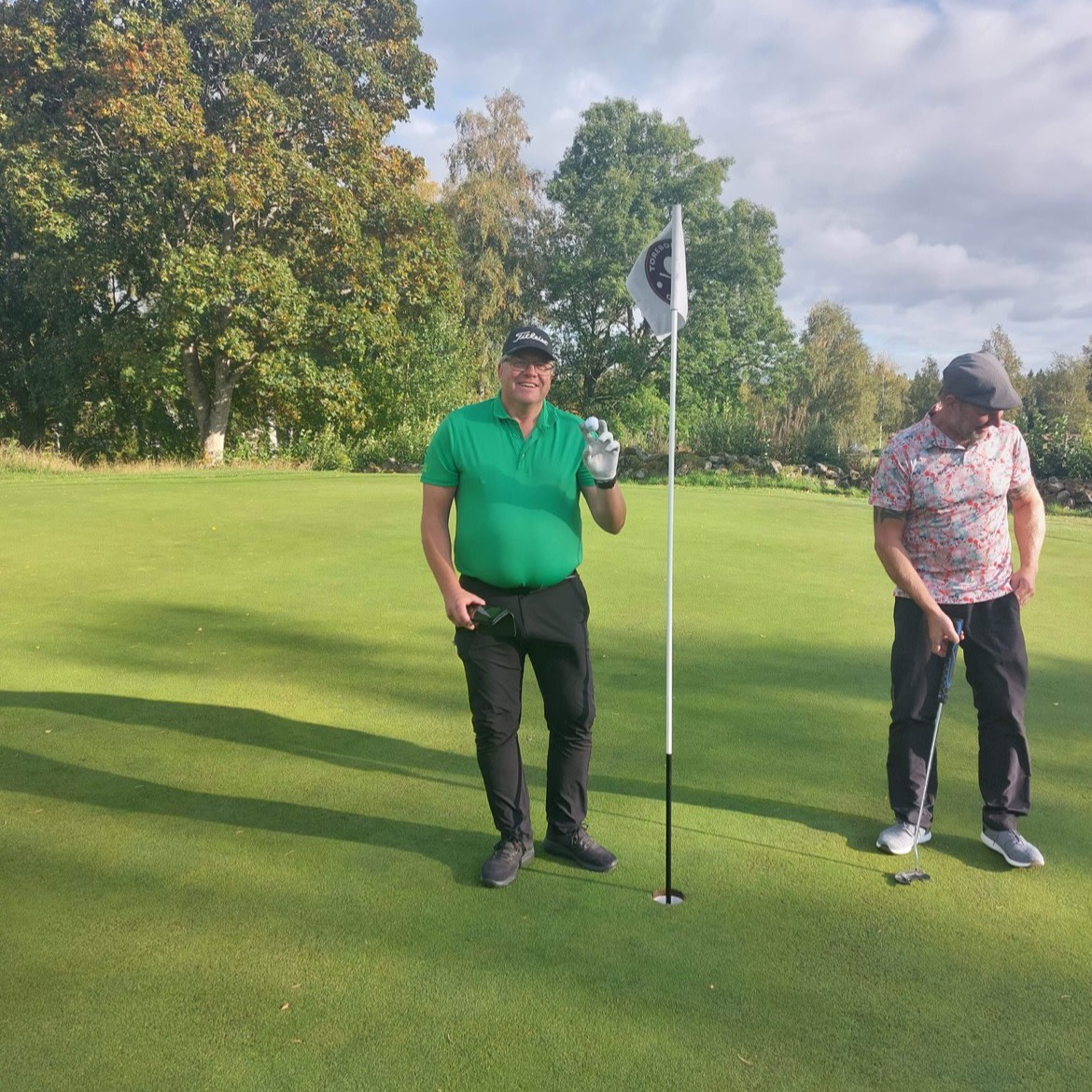 Två män står på en golfgreen, en man i grön tröja håller upp en golfboll bredvid flaggstången, den andra mannen lutar sig över sin putter.