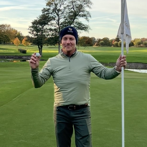 Man holder en golfball i den ene hånden og flaggstangen i den andre på en golfbane med grønt gress og trær i bakgrunnen.