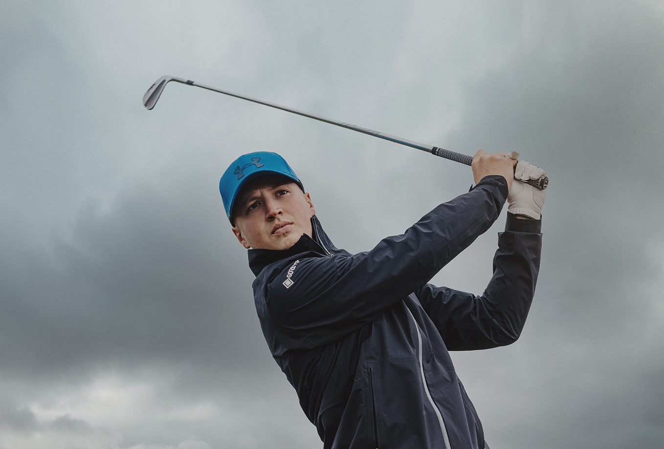 Mies sinisessä lippiksessä ja takissa heilauttaa golfmailaa ulkona pilvisen taivaan alla.