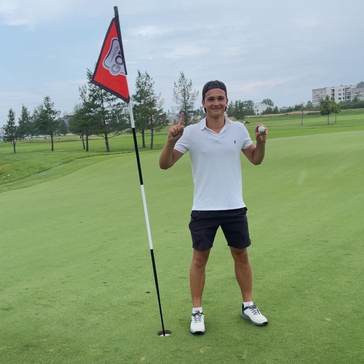 Man i vit pikétröja och svarta shorts står bredvid golfhål, håller upp en golfboll och pekar uppåt på en golfbana.