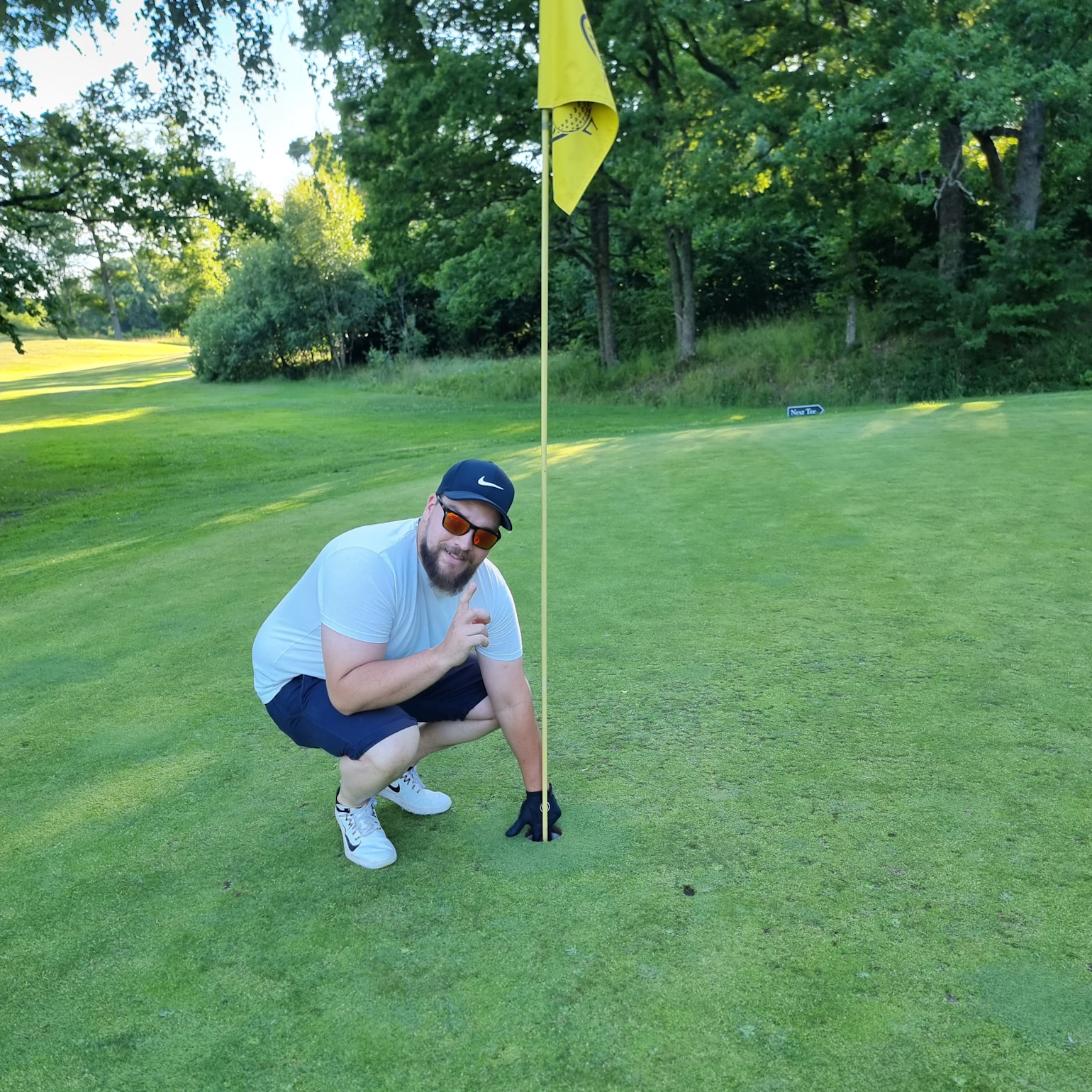 Mand med kasket og solbriller sidder på hug ved et golfhul på greenen, peger på hullet ved siden af flagstangen, omgivet af grønne omgivelser.