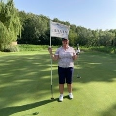 En person står på en golfbane og holder i en flagstang, omgivet af grønne områder og træer under en klar himmel.