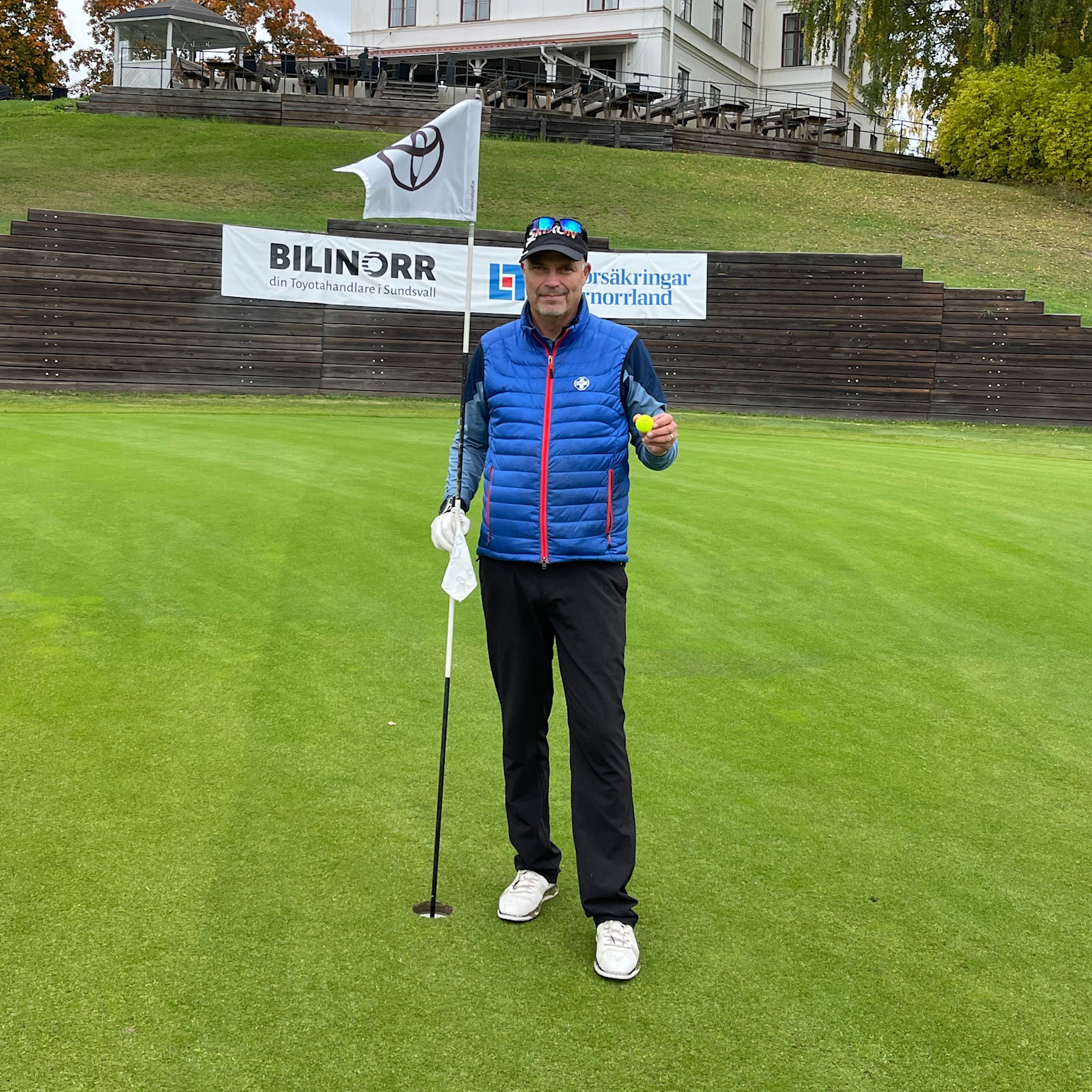 Man holder en golfbold og flagstang på en golfgreen, med reklameskilte og klubhus i baggrunden.