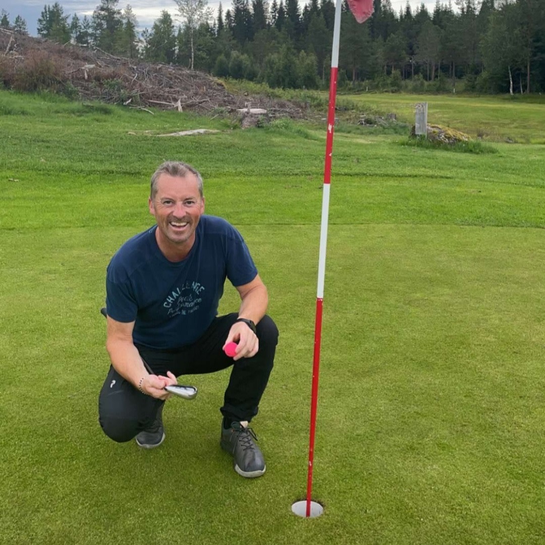 Smilende mand sætter sig på hug ved siden af et golfhul med flagstang, holder en lyserød golfbold på en grøn golfbane med skov i baggrunden.