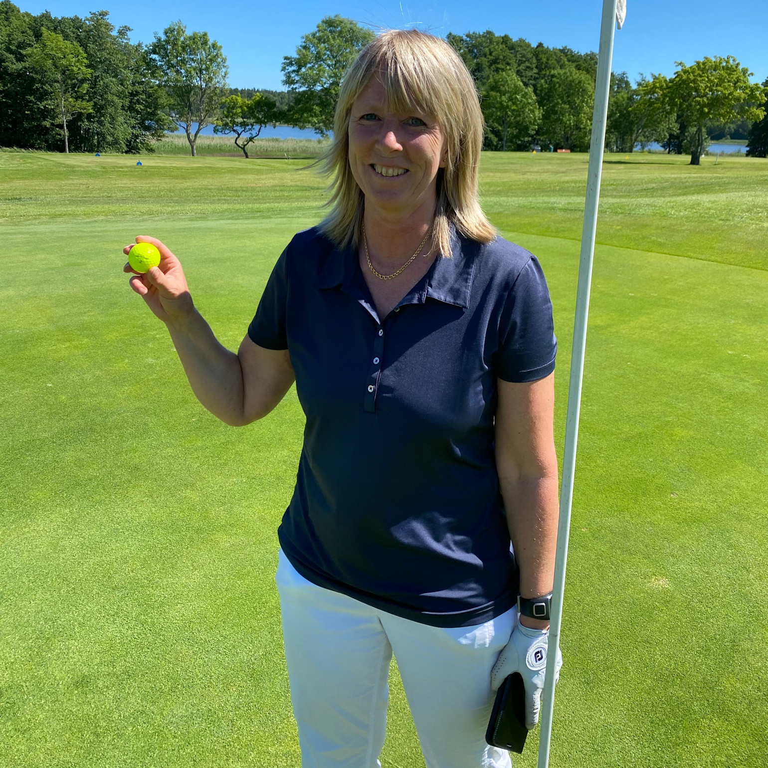 Kvinde i marineblå poloshirt og hvide bukser holder en gul golfbold ved siden af flagstangen på en golfbane i solskinsvejr.