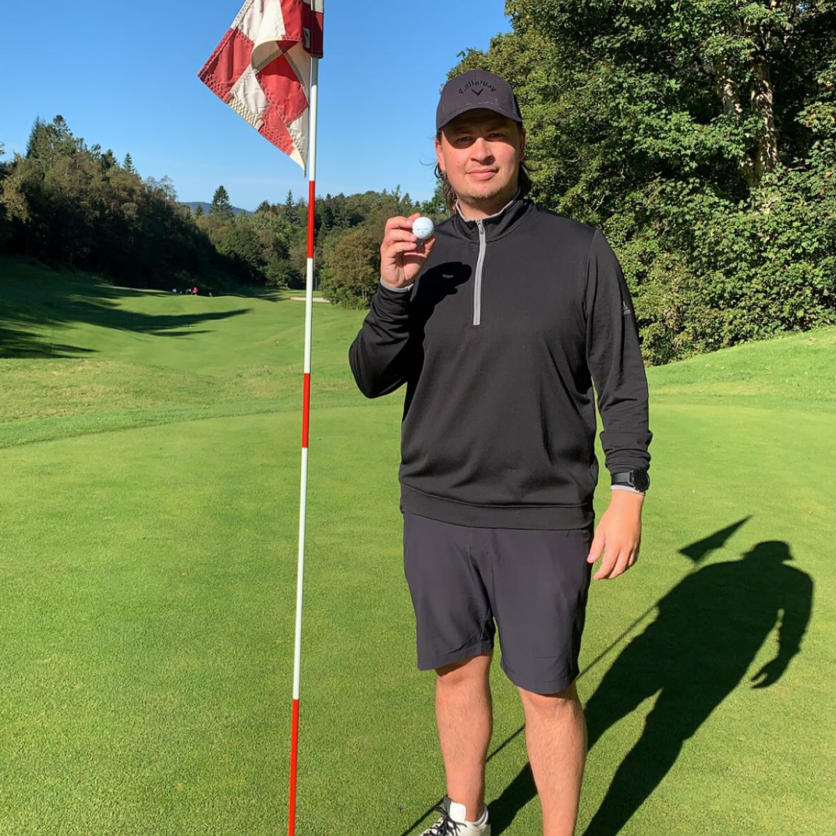Mand i sort golfbeklædning holder en golfbold op ved siden af en flagstang på en solrig golfbane med grønne omgivelser i baggrunden.