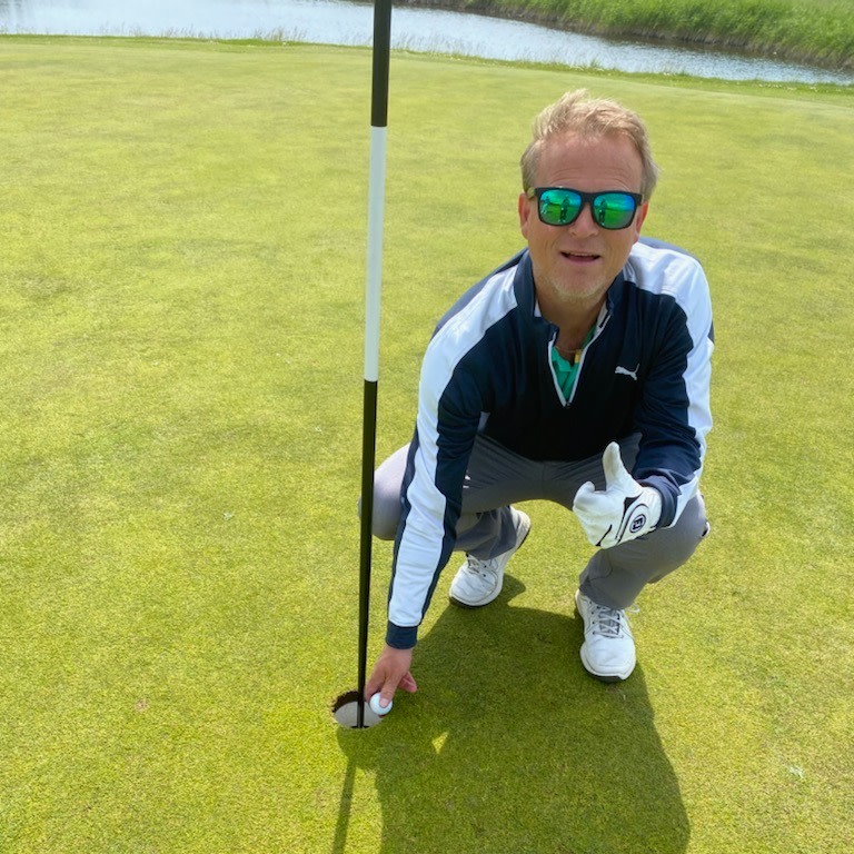 Mand i golfbeklædning samler en golfbold op fra hullet og giver tommelfingeren op på en golfbane nær vandet.