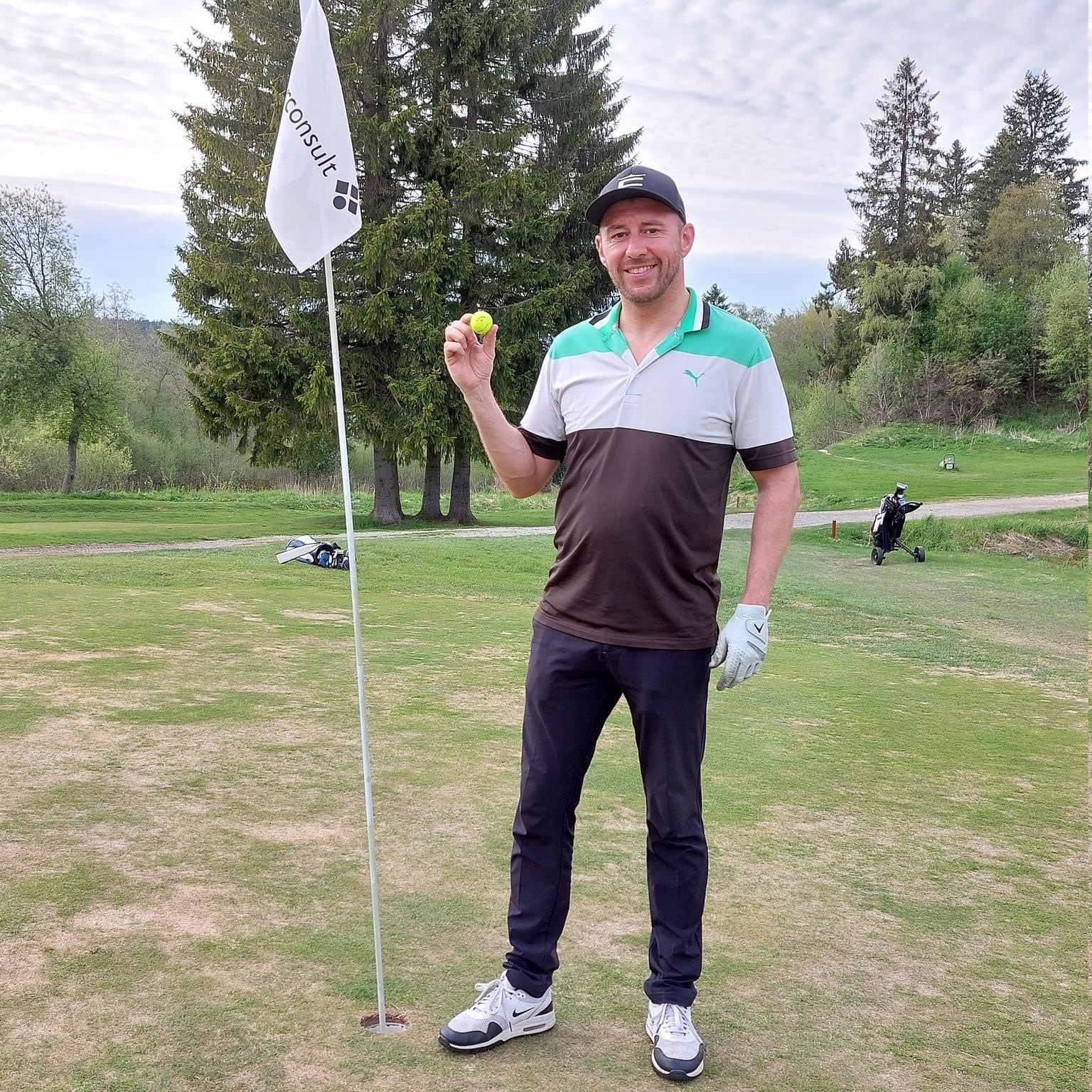 Man i golfkläder håller en gul golfboll bredvid en flaggstång på en golfbana med träd i bakgrunden.
