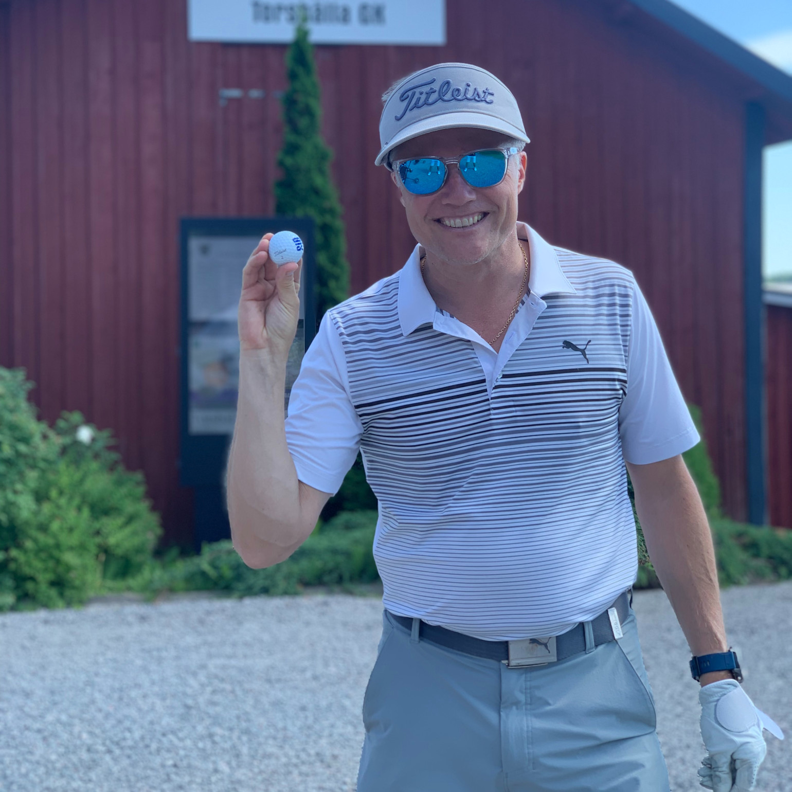 Mand i golfbeklædning holder en golfbold og smiler udendørs foran en rød bygning med grøn beplantning i baggrunden.