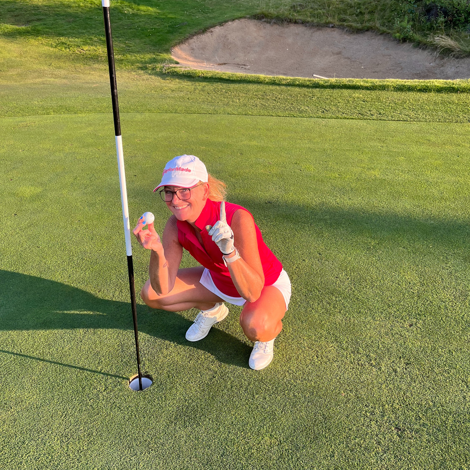 Kvinde i rød trøje og hvid kasket huker sig ved golfhul, holder en golfbold og viser én finger, golfbane i baggrunden.