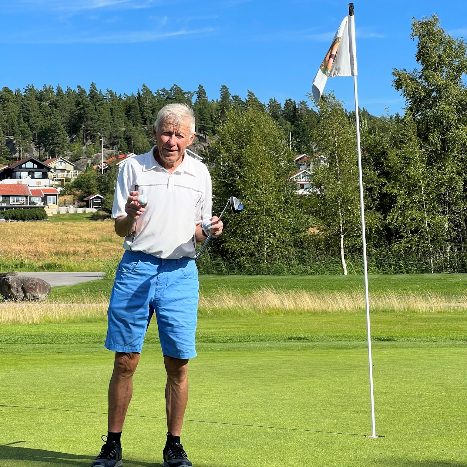 Ældre mand står på en golfgreen, holder en golfkølle og en golfbold ved siden af et hul med flag, med træer og huse i baggrunden.