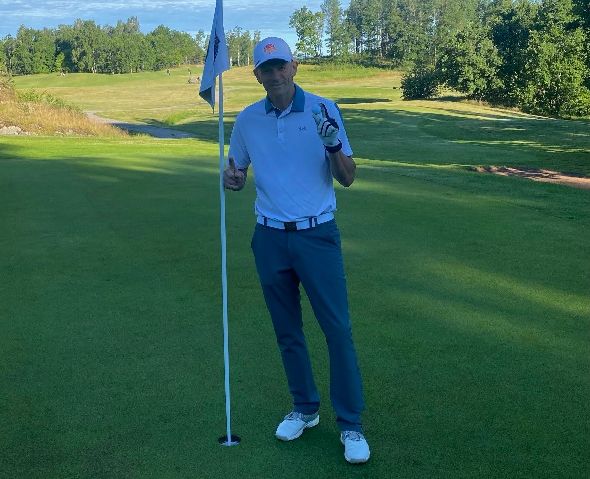 En mand i golfbeklædning står ved et hul på en golfbane, holder en golfbold og giver tommelfingeren op, omgivet af grønne områder og træer.