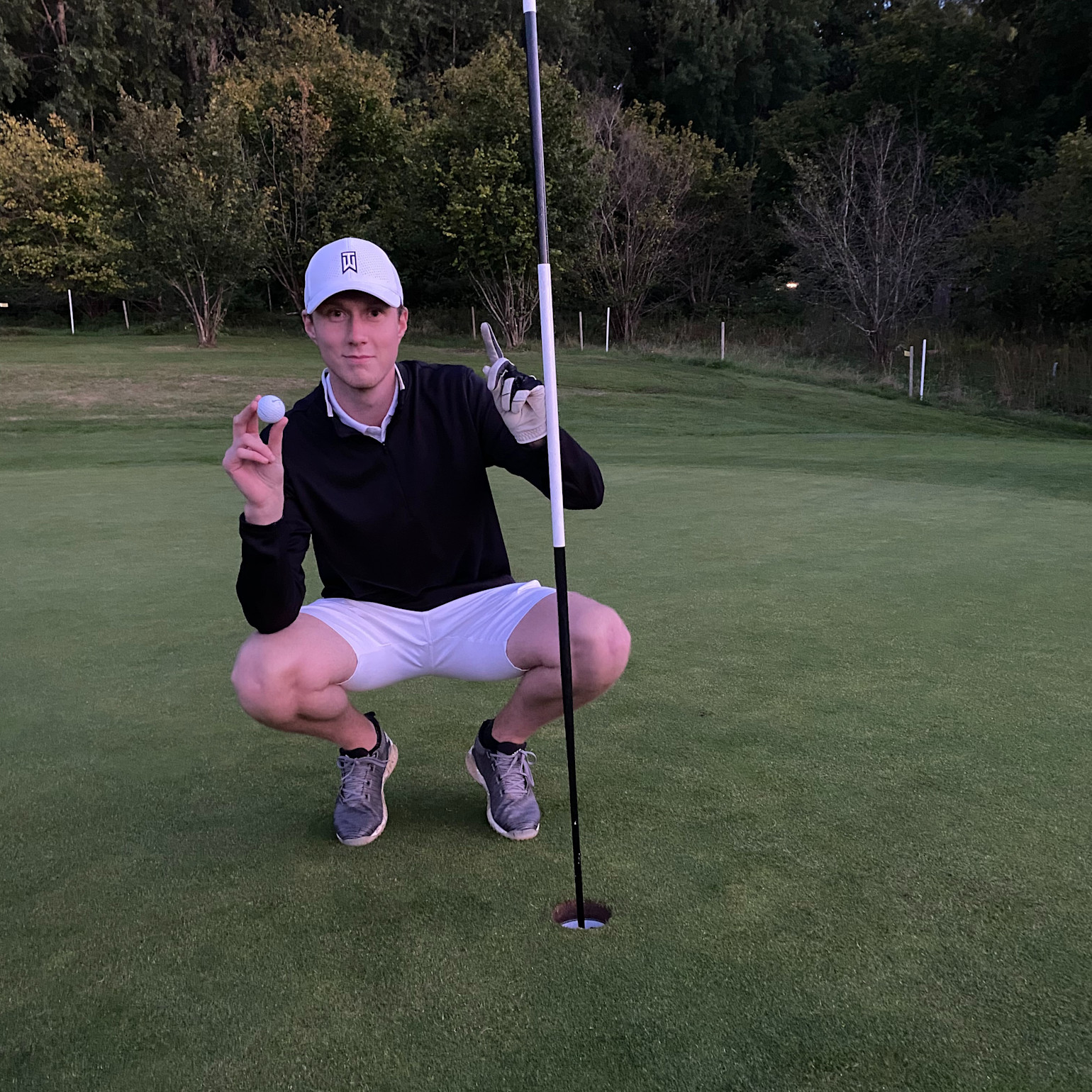 En mand iført golfbeklædning sidder på hug ved et golfhul, holder en golfbold op og peger mod flagstangen på en golfbane.