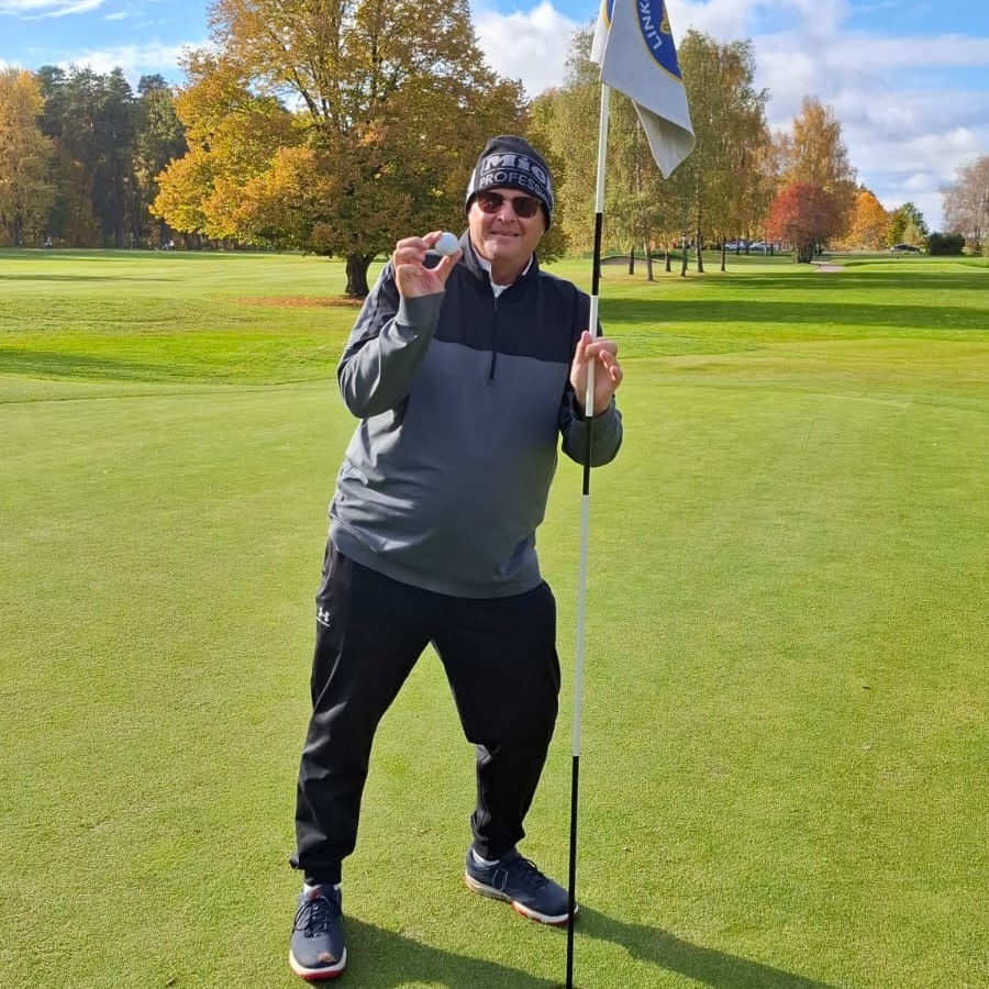Man holder en golfball og står ved siden av en flaggstang på en golfgreen på en solrik høstdag med trær i bakgrunnen.
