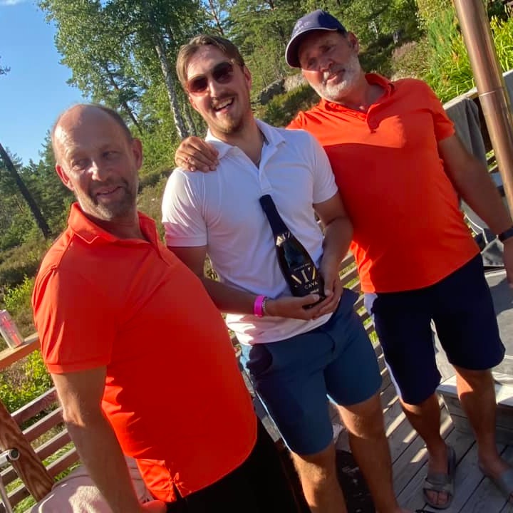 Tre män poserar utomhus, två i orange pikétröjor och en i vit, där den i mitten håller en vinflaska och ler mot kameran.