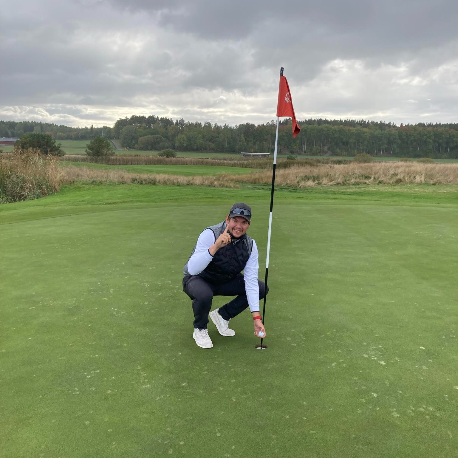 Mies juhlii hole-in-one -lyöntiään poseeraamalla golfreiän ja lipun vieressä golfkentällä, jonka taustalla näkyy vehreyttä ja pilvinen taivas.