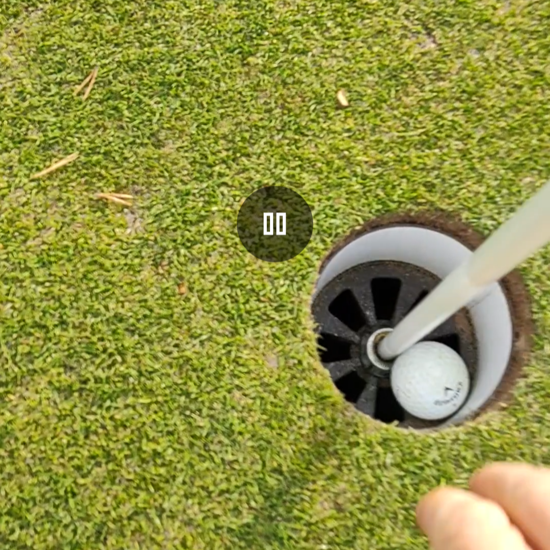 En golfbold ligger i hullet ved siden af flagstangen på en puttinggreen, med en hånd der rækker ud mod bolden.