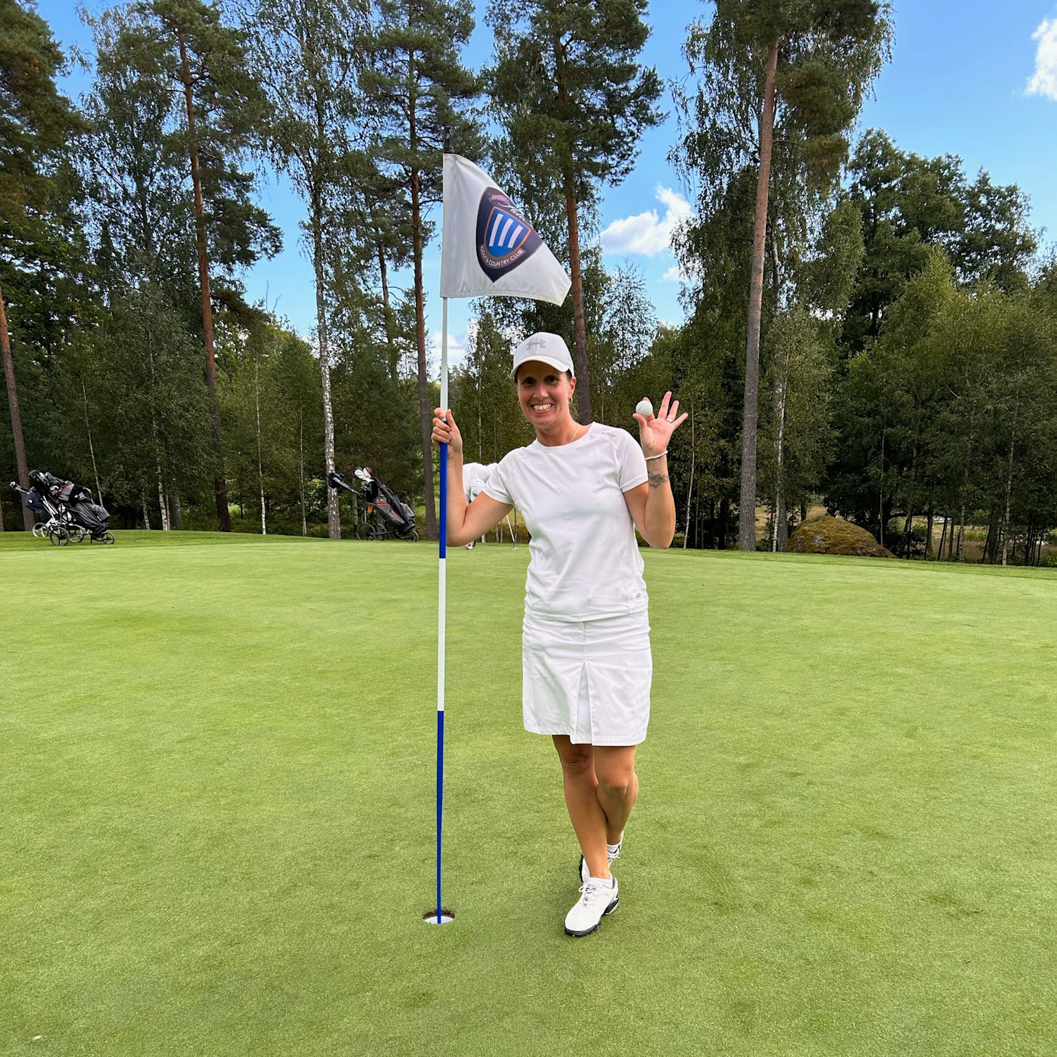 Kvinna i vit golfklädsel håller i flaggstång och golfboll på green, ler och vinkar, omgiven av träd och golfbagar.