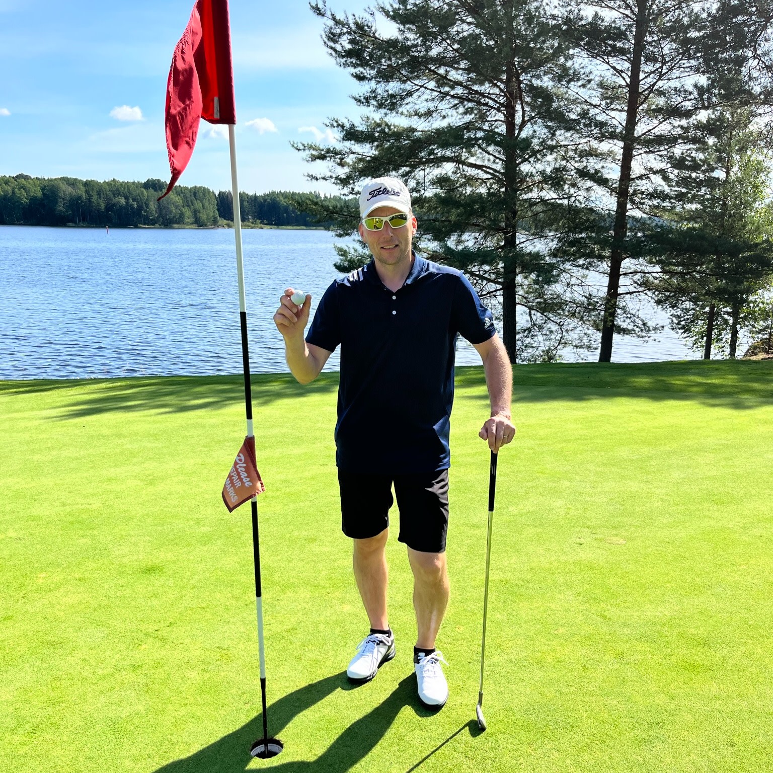 Mand iført golfbeklædning holder en golfbold og en golfkølle ved siden af et hul med flag på en golfbane ved en sø og nogle træer.